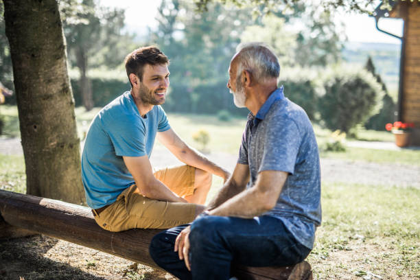 Happy adult son talking to his father in the back yard. Happy adult son communicating with his senior father while relaxing on a bench in nature. father in law stock pictures, royalty-free photos & images
