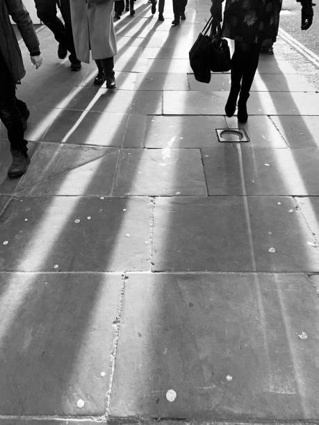 ​Busy city street with people walking and shadows cast on the pavement.  People only from waist down.