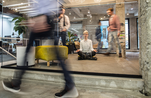 Young businesswoman exercising Yoga in Lotus position on the floor at casual office while their colleagues are walking in blurred motion. The view is through the glass.