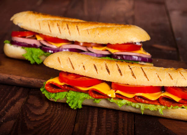 dos sándwiches de baguette con salami, jamón, queso, lechuga, tomate y cebolla en una tabla de cortar. - sandwich submarine delicatessen salami fotografías e imágenes de stock