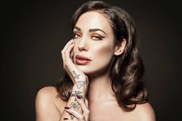 Classy Sexy Woman With Make-up and Tattoos Very sensual and beautiful woman with creative make-up and an elegant hairdo. Leaning on her hand, bare shoulders, no clothes. Several religious tattoos on her arm and polished nails. Head and shoulders, looking at camera. Model Dorien Rose, Dutch Playmate. cross shoulder tattoos stock pictures, royalty-free photos & images