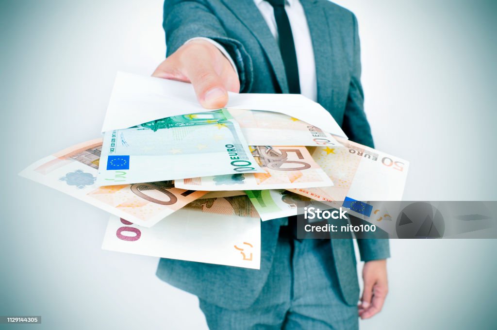 man giving an envelope full of euro bills a young caucasian man wearing a gray suit gives an envelope full of euro bills to the observer Currency Stock Photo
