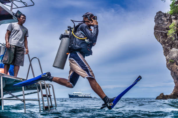 Bida Nok, Phi Phi Islands, Andaman sea, Thailand - May 31 2018:  One Asian man leaps off a boat into the Andaman sea.  A scuba diver demonstrating perfectly what is known as the ‘giant stride entry’.  Regulator in his mouth and wearing scuba equipment in place, he holds his mask in place.  Representative of the Millennial generation of eco tourists who spend much of their finances on environmentally friendly activities and travel.