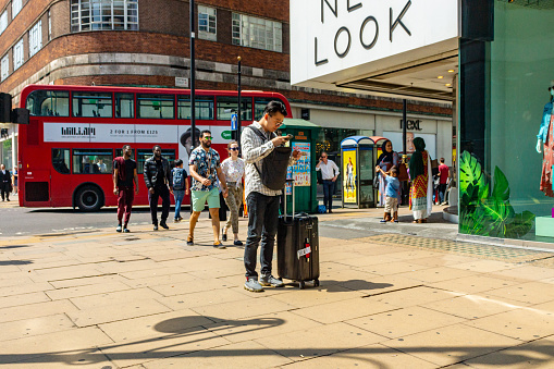 Crowds of shoppers on Regent Street in Central London, UK in the summer of 2018. a young Asian tourist looks lost outside New Look whilst he reads his mobile phone directions.