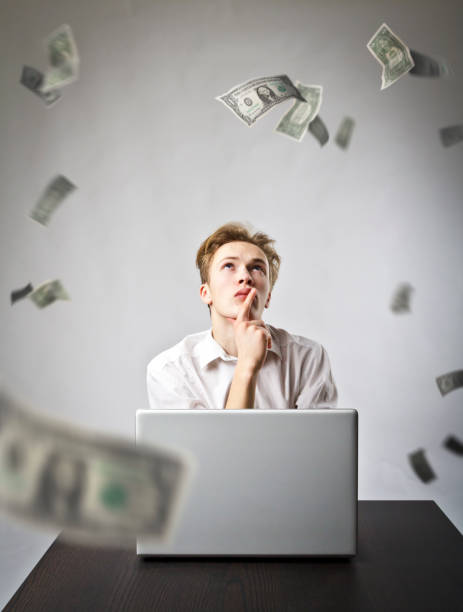 istock Young man using a laptop to browse the net. Young worker and falling dollar banknotes. Currency and job concept. 1129151007