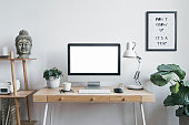 Stylish scandinavian interior of home creative desk with mock up computer screen, plants, bookstand and buddha figure. Minimalistic space for work, hobby and listen music. Freelancer zone.