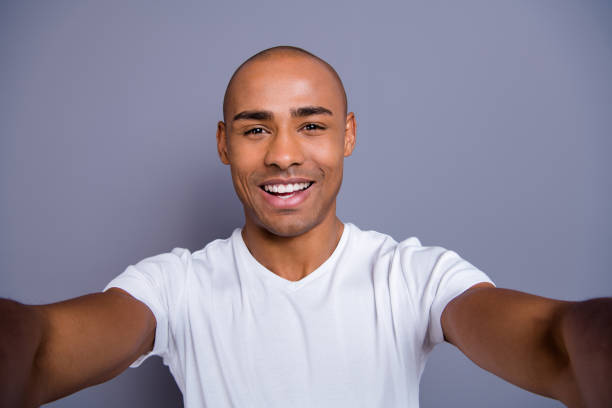 cerrar piel oscuro masculino foto él él su teléfono brazos cabeza calva macho hacer tomar autorretratos alegres con blanco camiseta equipo ropa aislado fondo gris - video conference camera fotografías e imágenes de stock