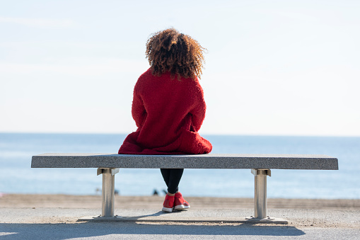 Rear view of young curly woman wearing red denim jacket sitting on a bench while looking away to horizon over sea