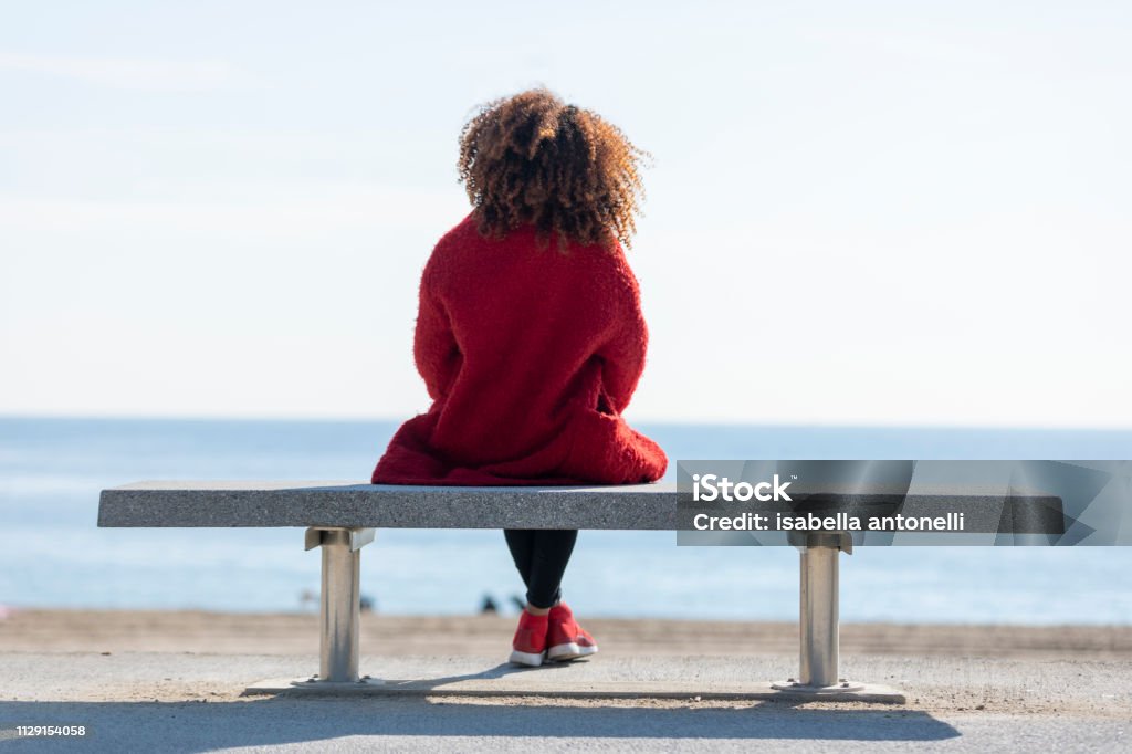 Rear view of a young curly woman wearing red denim jacket sitting on a bench while looking away to horizon over sea Rear view of young curly woman wearing red denim jacket sitting on a bench while looking away to horizon over sea Rear View Stock Photo