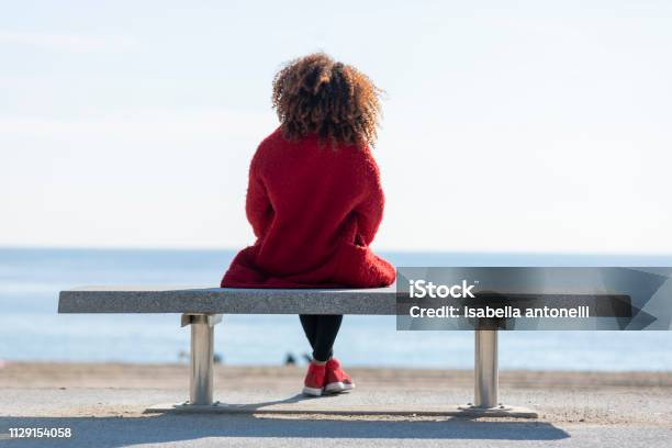 Rear View Of A Young Curly Woman Wearing Red Denim Jacket Sitting On A Bench While Looking Away To Horizon Over Sea Stock Photo - Download Image Now