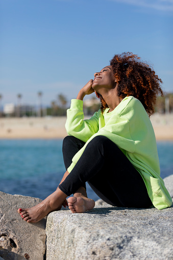 Side view of a beautiful curly afro woman sitting on breakwater rocks laughing while resting hand under chin eyes closed outdoors