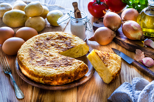 Spanish cuisine: High angle view of a rustic wooden kitchen table filled with traditional spanish tortilla ingredients. A prepared spanish tortilla with a slice is at the left surrounded by ingredients for cooking tortilla like eggs, potatoes, onion, garlic, parsley, salt and olive oil. Predominant colors are yellow and brown. DSRL studio photo taken with Canon EOS 5D Mk II and Canon EF 100mm f/2.8L Macro IS USM.