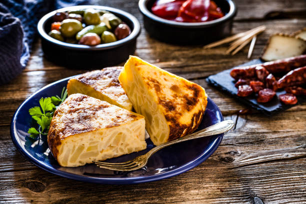 closeup of a plate with tortilla de patatas, spanish omelet, served as tapas, a glass with tinto de verano, bread and a bowl with olives on a colorful blue wooden table
