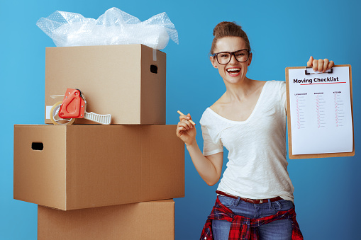 happy modern woman in white t-shirt near cardboard box showing moving checklist against blue background