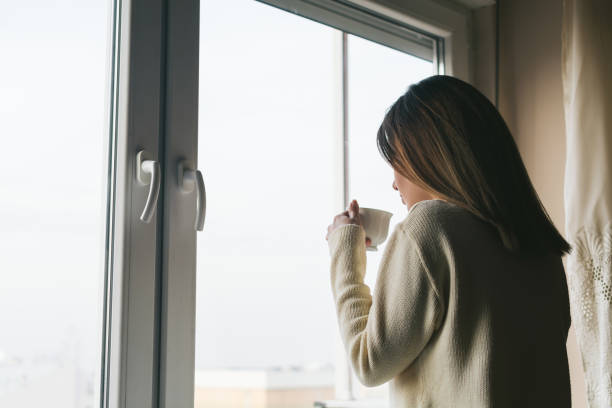 istock Young woman drinking coffee at home and looking through the window 1129178287