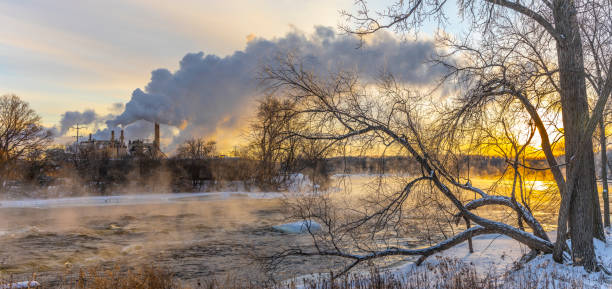 Steaming river coats trees with ice in subzero cold Winter sunrise, factory emissions in background.