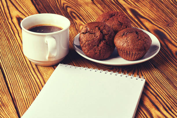 Several chocolate muffins with dark pastry with a cup of coffee and a notepad on a wooden table.