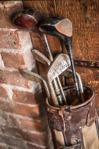 Old-fashion wooden and iron golf clubs in a rustic golf bag resting against a brick wall.