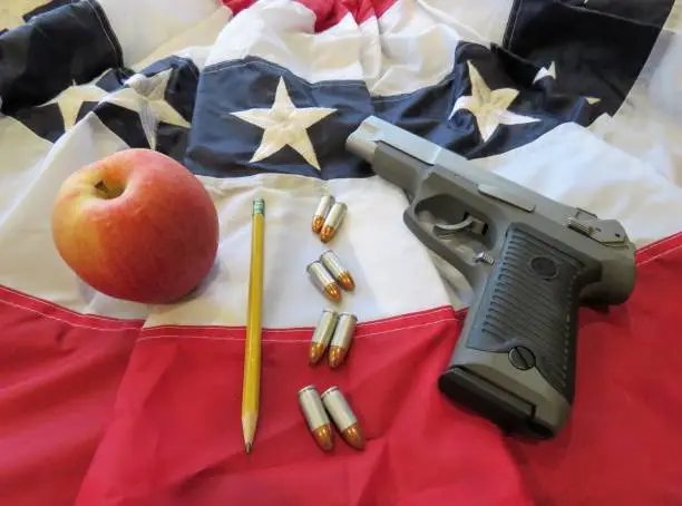 school shootings & gun control debate symbolized by an apple, pencil, bullets, gun on a US flag bunting