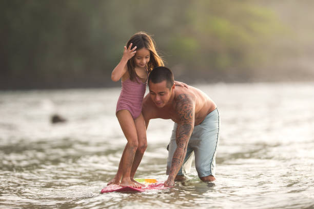 il papà eurasiatico insegna alla sua piccola ragazza come navigare - surfing beach family father foto e immagini stock