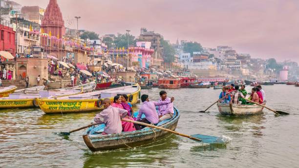 indian tourists being rowed along the ganges river, passing the waterfront ghats filled with hindu pilgrims who bathe in the river. - morning river ganges river varanasi imagens e fotografias de stock