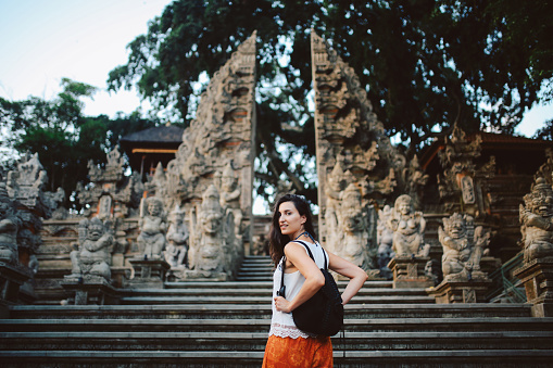 Image of a young woman walking by the temple in Bali, Indonesia. Decorated entrance to the old Balinese temple - shrine. She is holding a backpack and wearing casual, comfortable colorful yogi pants.