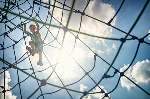 Portrait of little boy climbing in the playground. The boy aged 8 is smiling down at the camera from the climbing web. The sun and blue sky visible in the background.
Nikon D850