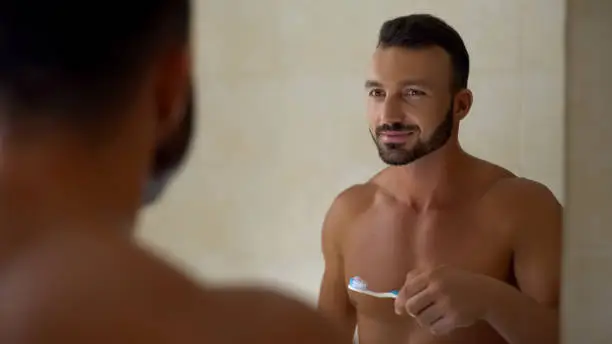 Man holding toothbrush with toothpaste looking in mirror at bathroom, dentalcare