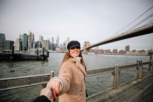From the perspective of a photographer, a woman poses for the camera by Brooklyn Bridge, New York City.