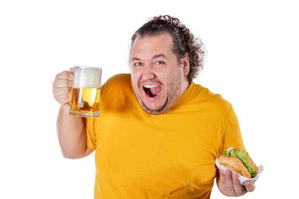 istock Funny fat man eating burger and drinking alcohol beverage on white background 1129329595