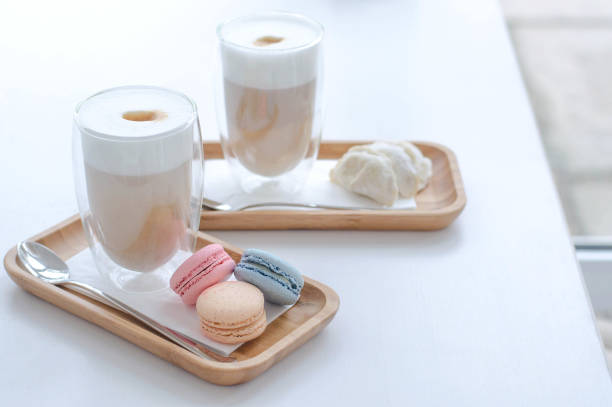 Two latte cups with macaroons and cookies. Two cups with a latte and a row of colorful macaroons and cookies on a wooden stand with a spoon. cafe transparent glass table stock pictures, royalty-free photos & images