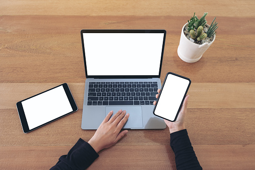 Top view mockup image of a hands holding a blank white screen mobile phone and laptop with black tablet on wooden table in office