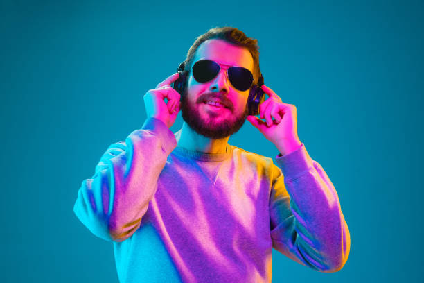 Enjoying his favorite music. Enjoying his favorite music. Happy young stylish man in hat and sunglasses with headphones listening and smiling while standing against blue neon background dance  electronic music photos stock pictures, royalty-free photos & images