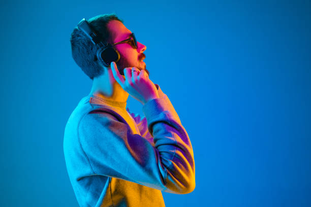 Enjoying his favorite music. Enjoying his favorite music. Happy young stylish man in sunglasses with headphones listening and smiling while standing against blue neon background dance  electronic music photos stock pictures, royalty-free photos & images