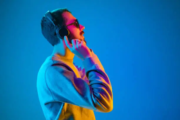 Enjoying his favorite music. Happy young stylish man in sunglasses with headphones listening and smiling while standing against blue neon background
