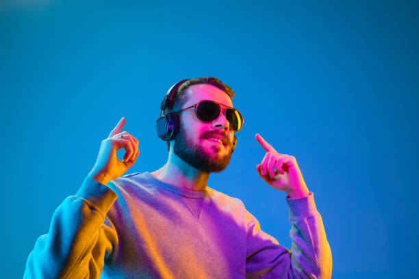 Enjoying his favorite music. Enjoying his favorite music. Happy young stylish man in hat and sunglasses with headphones listening and smiling while standing against blue neon background dance  electronic music photos stock pictures, royalty-free photos & images