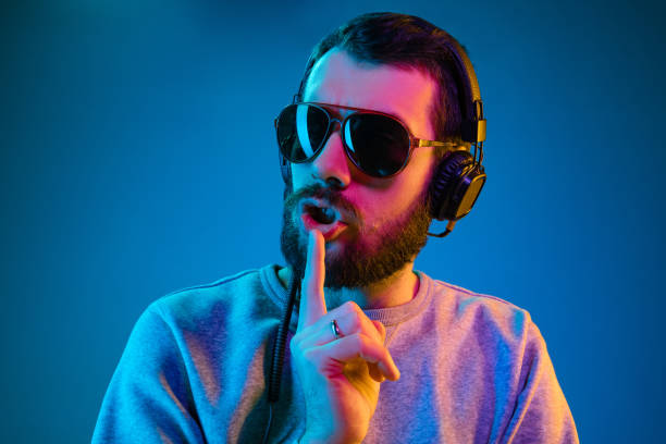 Enjoying his favorite music. Enjoying his favorite music. Happy young stylish man in sunglasses with headphones listening sound and calling for silence while standing against blue neon background dance  electronic music photos stock pictures, royalty-free photos & images