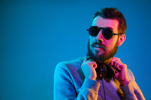 Enjoying his favorite music. Happy young stylish man in sunglasses with headphones smiling while standing against blue neon background