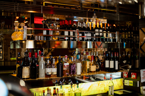 Front view of behind the bar counter in a restaurant. There are bottles of wine and various spirits on shelves.