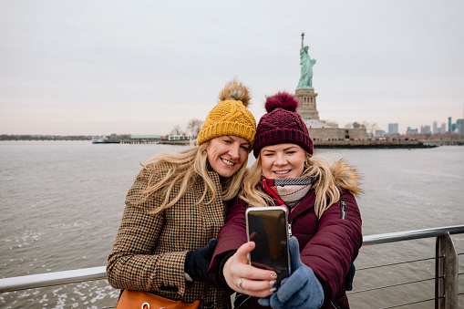 Two women taking a selfie on a tour boat in New York City in front of the Statue of Liberty.