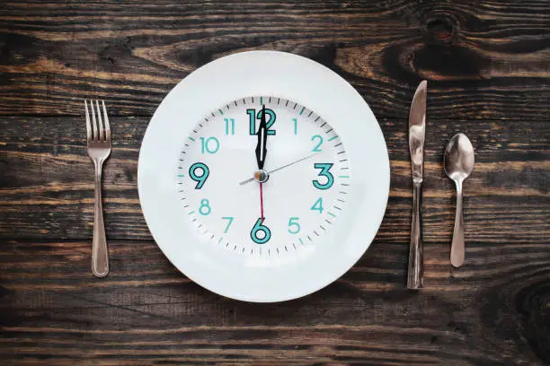 Twelve hour intermittent fasting time concept with clock on plate over a rustic wooden table / background. Top view.