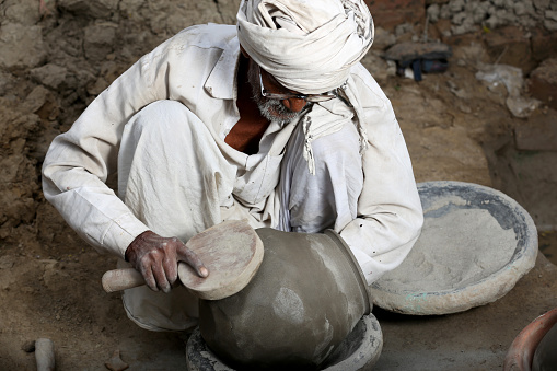 Traditional Indian Potter Sitting wearing white Kurta-Pajama & Turban which is Traditional Dress for Men in North India. He is hitting the Clay Pots to giving it Proper Round Shape near brick wall .