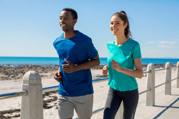 Smiling multi-ethnic athletes jogging on promenade by railing against sky. Young couple are running at beach on sunny day. They are in sports clothing.