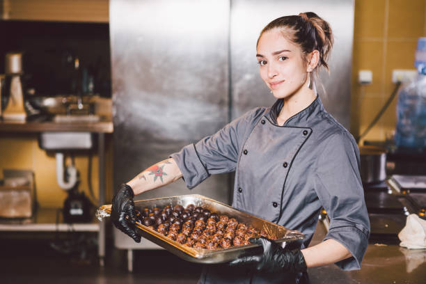 thema beruf und kochen kuchen. kaukasische mädchen mit tattoo eines konditors in küche des restaurants vorbereitung runde schokolade süßigkeiten handgemachte trüffel in schwarzen handschuhen und uniform - konditor stock-fotos und bilder