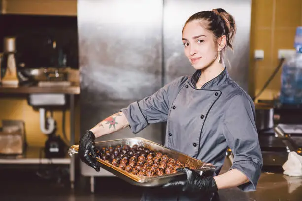 Subject profession and cooking pastry. young Caucasian woman with tattoo of pastry chef in kitchen of restaurant preparing round chocolate candies handmade truffle in black gloves and uniform.