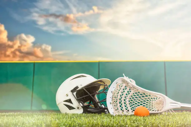 A Lacrosse stick with an orange ball and a B&W helmet sitting on a synthetic grass turf with green padded wall around the sports field and sunbeams from the setting sun.