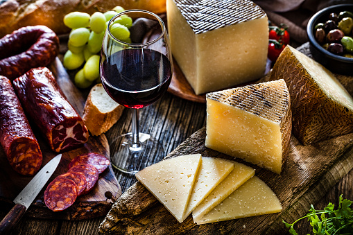 Spanish food: Manchego cheese slices and pieces in a cutting board shot on dark rustic wooden table. A red wine glass is beside the Manchego pieces. Spanish chorizo is visible at the left. A bowl filled with olives, bread and green grapes complete the composition. Predominant colors are brown and yellow. Low key DSRL studio photo taken with Canon EOS 5D Mk II and Canon EF 100mm f/2.8L Macro IS USM.