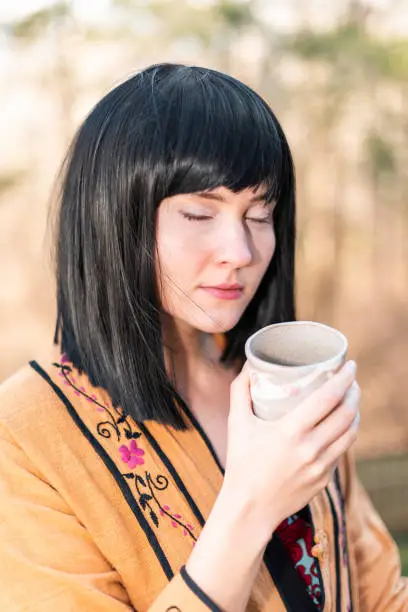 Woman girl black hair holding matcha green tea cup face serious closeup drinking outside in backyard garden with closed eyes