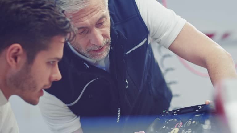 Mature coach explaining controls of steering wheel to racer. Males are having discussion in auto repair shop. They are preparing for open-wheel single-seater racing car competition.