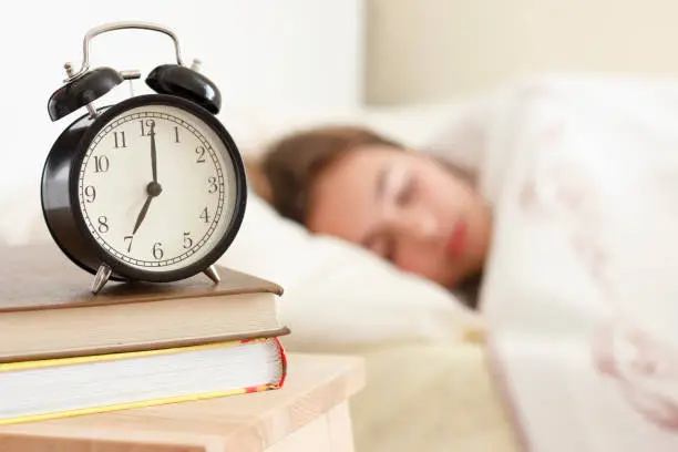 Teenager girl sleeping in a white bed. Alarm clock in the foreground on pile of books